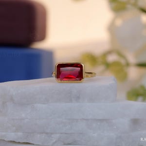 May include: A gold ring featuring a rectangular, deep red gemstone. The ring is displayed on a white, textured surface. The gemstone is the focal point, with its rich colour enhanced by the gold setting. The ring's design is simple, highlighting the gemstone's beauty.