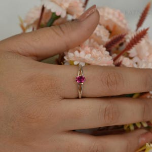 May include: A gold ring with a vibrant oval-cut pink gemstone. The ring's band splits into two delicate prongs that cradle the gemstone. The ring is displayed on a hand, with a soft floral arrangement in the background.
