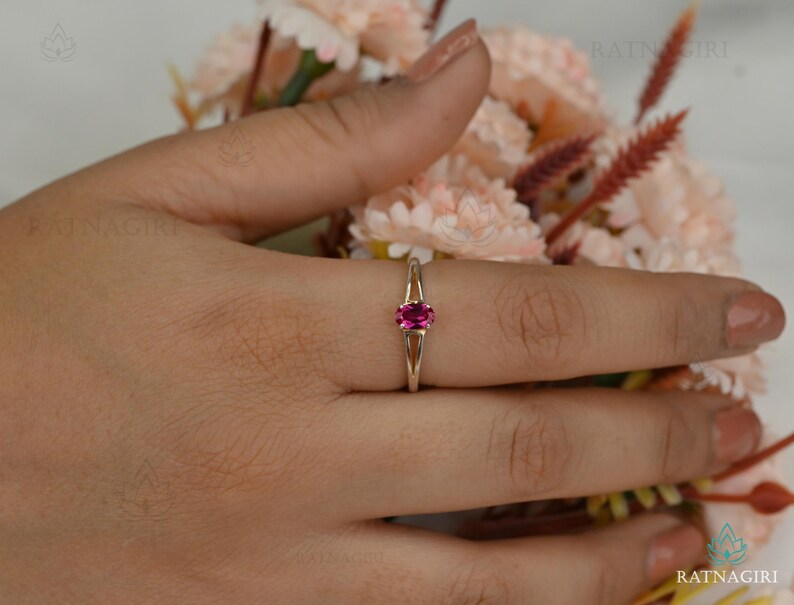 May include: A gold ring with a vibrant oval-cut pink gemstone. The ring's band splits into two delicate prongs that cradle the gemstone. The ring is displayed on a hand, with a soft floral arrangement in the background.