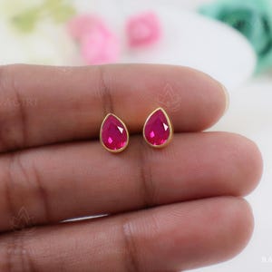 May include: A pair of gold-colored stud earrings, each featuring a teardrop-shaped, vibrant pink gemstone. The earrings are set in a simple, elegant design, showcasing the bright color of the stones. The earrings are displayed against a soft, neutral background.