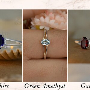 May include: Three silver rings with oval gemstones: a deep blue sapphire, a light blue green amethyst, and a dark red garnet. The rings are displayed against neutral backgrounds. Text at the bottom reads "Sapphire", "Green Amethyst", and "Garnet".
