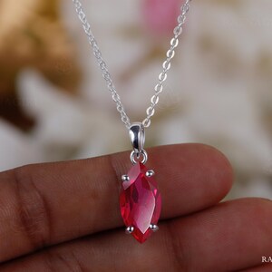 May include: A silver necklace featuring a marquise-cut, ruby-red gemstone pendant. The pendant is set in a silver bezel and suspended from a delicate silver chain. The gemstone is a vibrant red colour.