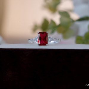 Könnte beinhalten: Ein silberner Ring mit einem großen, rechteckigen, tiefroten Edelstein als Mittelpunkt. Zwei kleinere, klare Edelsteine flankieren den Hauptstein. Der Ring liegt auf einer weißen Oberfläche, mit einem verschwommenen Hintergrund aus Grün.