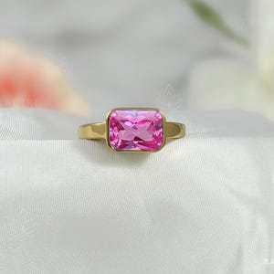 Könnte beinhalten: Ein goldener Ring mit einem rechteckigen, leuchtend rosa Edelstein. Der Ring hat ein schlichtes, elegantes Design mit einem glatten, polierten Band. Der Edelstein ist in einer Lünettenfassung gefasst, die seine Farbe und Form hervorhebt. Der Ring wird auf einer weißen, strukturierten Oberfläche präsentiert.