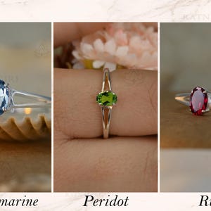 May include: Three rings with oval gemstones: aquamarine, peridot, and ruby. Each ring has a silver band. The aquamarine ring is on a seashell, the peridot ring is on a finger, and the ruby ring is on a neutral background. Text below reads "Aquamarine", "Peridot", and "Ruby".