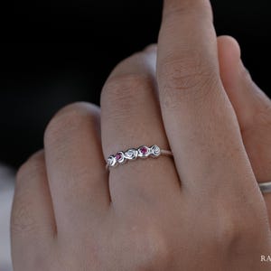 Peut inclure: Une bague en argent ornée d'une rangée de cinq pierres rondes alternées, comprenant trois pierres blanches transparentes et deux pierres rouge rosé éclatantes, serties sur un anneau métallique lisse. Le design présente une finition polie.