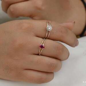 May include: Two rings on a finger. One ring features a small, round, white gemstone cluster. The other ring has a gold band with a small, oval, red gemstone. The rings are set against a neutral background.