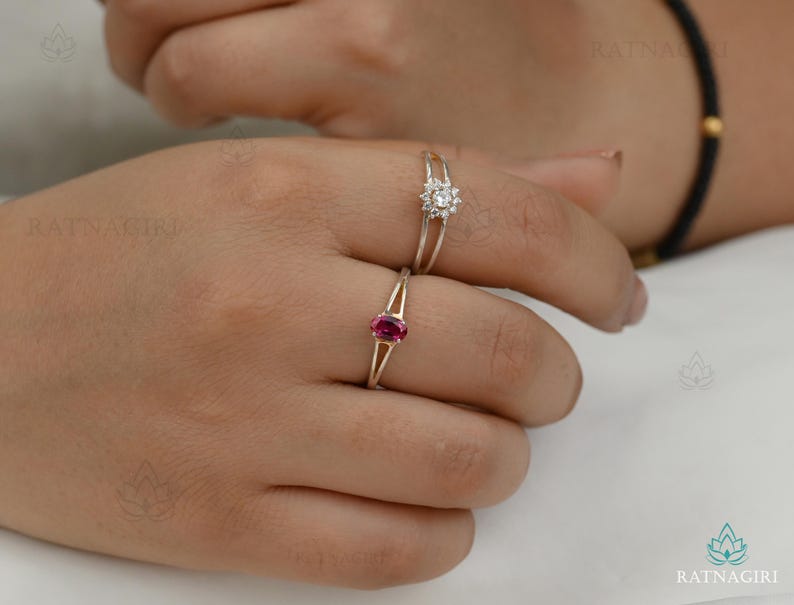 May include: Two rings on a finger. One ring features a small, round, white gemstone cluster. The other ring has a gold band with a small, oval, red gemstone. The rings are set against a neutral background.