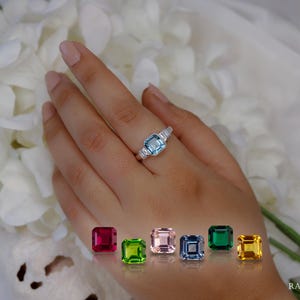 May include: A silver ring featuring a large, light blue gemstone and smaller white stones. The ring is on a hand, with several colourful gemstones displayed below. White flowers are in the background. Image from Ratnagiri.