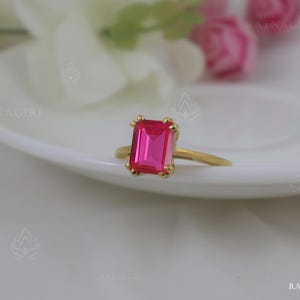 Könnte beinhalten: Ein goldener Ring mit einem leuchtend pinkfarbenen quadratischen Edelstein.