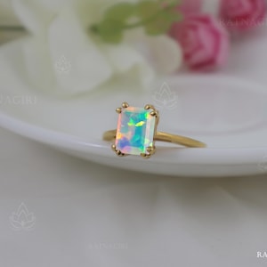 Puede incluir: Un anillo de oro con una gran piedra de ópalo rectangular e iridiscente. El anillo está colocado sobre una superficie blanca.