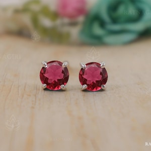 May include: A pair of stud earrings featuring round, deep red gemstones set in silver-colored metal. The earrings are displayed on a light wooden surface, with a blurred floral background. The gemstones have a faceted appearance, catching the light.