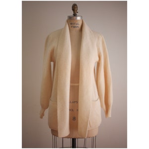 May include: A cream-colored knitted cardigan with a wide collar and two pockets. The cardigan is on a dress form.