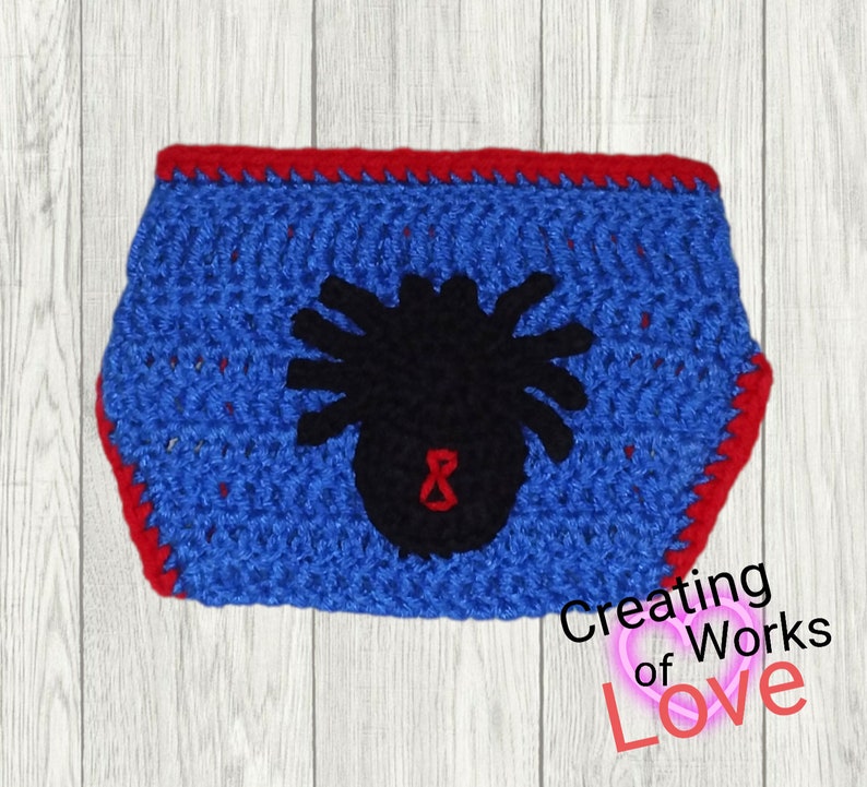 Puede incluir: Pa&ntilde;al de beb&eacute; de crochet azul con ribete rojo y una aplicaci&oacute;n de ara&ntilde;a negra con un ocho rojo en el centro.