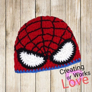 Puede incluir: Sombrero de punto rojo y negro con un dise&ntilde;o de m&aacute;scara de Spider-Man blanca. El sombrero tiene un borde azul. El texto "Creating of Works of Love" es visible en la esquina inferior derecha de la imagen.