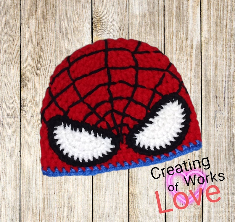 Puede incluir: Sombrero de punto rojo y negro con un dise&ntilde;o de m&aacute;scara de Spider-Man blanca. El sombrero tiene un borde azul. El texto "Creating of Works of Love" es visible en la esquina inferior derecha de la imagen.