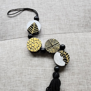 May include: A black and gold tassel keychain with a string of round wooden beads. Each bead is painted with a different geometric pattern in black and gold.