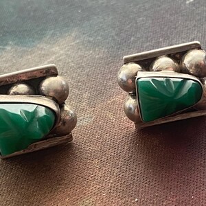May include: A pair of silver earrings featuring a central triangular green stone with a carved surface. The metal setting includes a horizontal bar at the top and bottom, accented by a row of three small silver spheres between the bars.