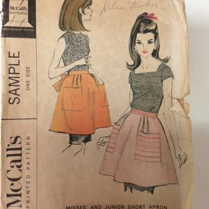 May include: Vintage McCall's printed pattern for Misses' and Junior short aprons. The illustration shows two apron designs, one in orange and the other in pink, with a gray top. The pattern is labeled "Sample" and "One Size".