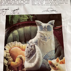 以下が含まれることがあります： 青いアクセントの布製猫人形が緑の椅子に座っています。猫は笑顔で、ハート型の枕や小さな猫の置物など、他の布製のアイテムに囲まれています。画像は縫製パターンからのものです。