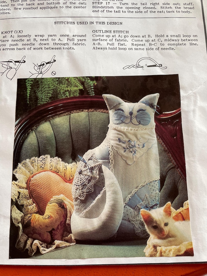 K&ouml;nnte beinhalten: Eine Stoffkatzenpuppe mit blauen Akzenten, die auf einem gr&uuml;nen Stuhl sitzt. Die Katze hat ein l&auml;chelndes Gesicht und ist von anderen Stoffartikeln umgeben, darunter ein herzf&ouml;rmiges Kissen und eine kleine Katzenfigur. Das Bild stammt aus einer N&auml;hanleitung.