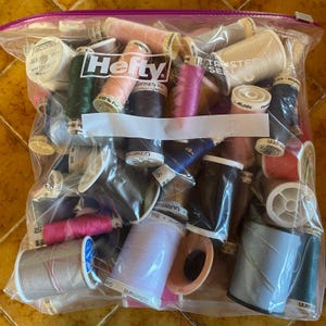 May include: A clear plastic Hefty storage bag filled with a variety of multi-colored sewing thread spools. The collection includes assorted sizes and shades of polyester thread, featuring brands like Gütermann and Mettler, visible through the transparent material with a purple zipper closure at the top.