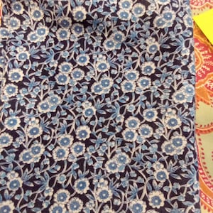 May include: A folded piece of fabric with a floral pattern. The design features small, light blue and white flowers with green stems and leaves against a dark navy blue background. The fabric has a soft, textured appearance.