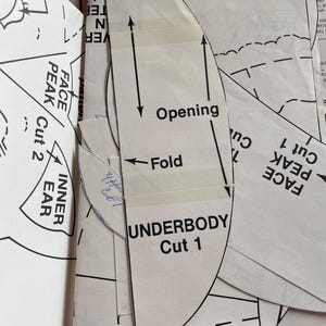 K&ouml;nnte beinhalten: Nahaufnahme von Schnittmustern mit Textetiketten wie "UNDERBODY Cut 1", "FACE PEAK Cut 2" und "INNER EAR". Die Schnittmuster sind wei&szlig; mit schwarzen Linien und Pfeilen und sind auf andere Schnittmusterb&ouml;gen geschichtet.