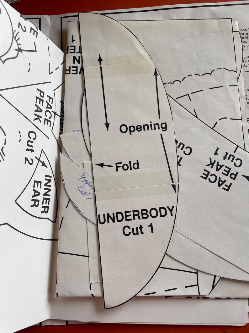 K&ouml;nnte beinhalten: Nahaufnahme von Schnittmustern mit Textetiketten wie "UNDERBODY Cut 1", "FACE PEAK Cut 2" und "INNER EAR". Die Schnittmuster sind wei&szlig; mit schwarzen Linien und Pfeilen und sind auf andere Schnittmusterb&ouml;gen geschichtet.