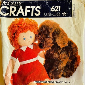 May include: Vintage McCall's Crafts pattern cover featuring a doll and a dog. The doll has red yarn hair and a red dress. The dog is brown and fluffy. The text reads "Annie and Friend Sandy Dolls". The cover is aged with wear.