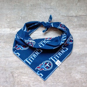 May include: Blue and white dog bandana featuring the Tennessee Titans logo and the words "Tennessee Titans" repeated on the bandana. The bandana has a white tag with the words "Doggy Fun Co."