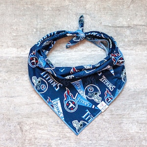 May include: Blue and white dog bandana with a repeated pattern of the Tennessee Titans logo and the word "Titans".