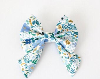 May include: A large fabric bow with a floral pattern in shades of blue, green, and gold on a white background. The bow is tied in the center and has two large loops and two tails. The fabric has a delicate, feminine design.