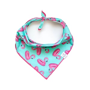 May include: A turquoise dog bandana with a pink flamingo floatie pattern and a pink border. The bandana is tied at the top. The tag on the bandana says "Pink Pups & Co."