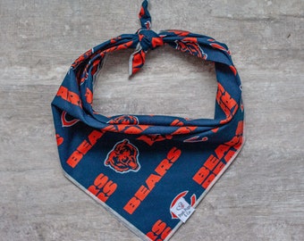 May include: A blue and orange dog bandana with the Chicago Bears logo and the text "Bears" repeated throughout. The bandana is tied in a knot at the top and has a small white tag with the text "Pawty Pup & Co."