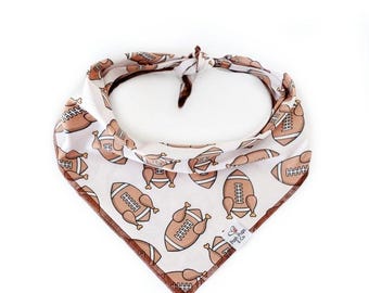 May include: White dog bandana with a brown football and turkey leg pattern. The bandana has a brown border and a small tag that says "Paw Prints & Co."