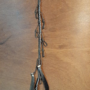 May include: A handmade horse hair tassel with a braided cord and a metal ring. The tassel features multiple knotted accents and dark gray horse hair strands, hanging against a wooden background.