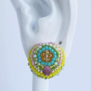 以下が含まれることがあります： 中央にオレンジ色の花模様をあしらい、その周囲をターコイズ、ライムグリーン、イエロー、ピンク、クリアホワイトの小さなガラスビーズで同心円状に囲んだ、密度が高く質感のある円形のスタッドピアス。