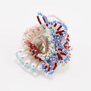 May include: A beaded brooch with a shell centerpiece, featuring a mix of white, blue, and red beads.