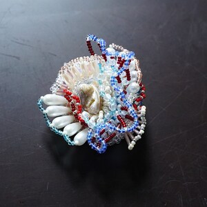 May include: A colorful beaded brooch with a shell centerpiece. The brooch features red, blue, white, and iridescent beads arranged in a swirling pattern.