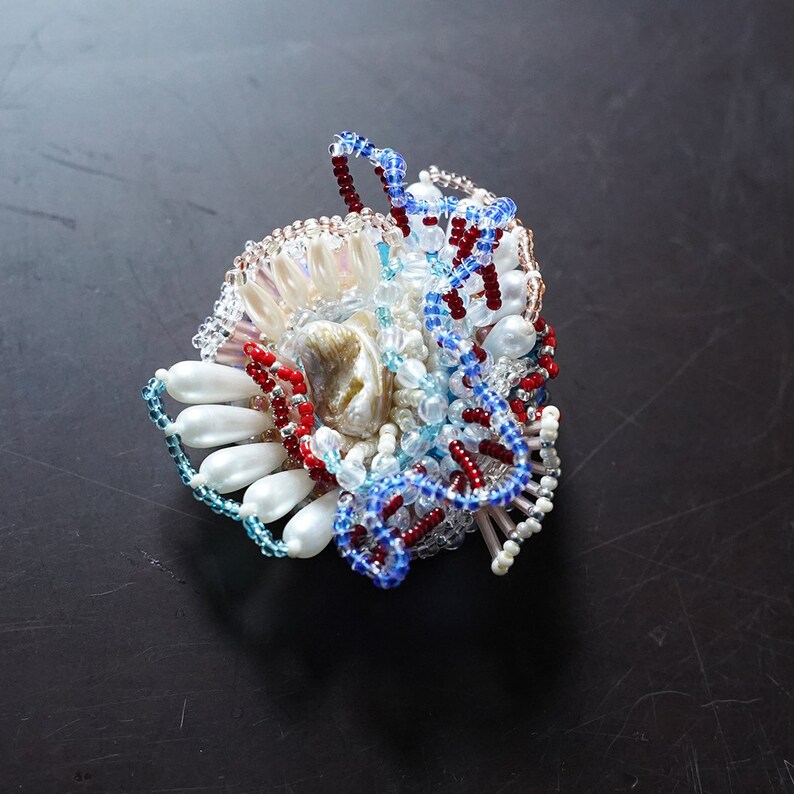 May include: A colorful beaded brooch with a shell centerpiece. The brooch features red, blue, white, and iridescent beads arranged in a swirling pattern.