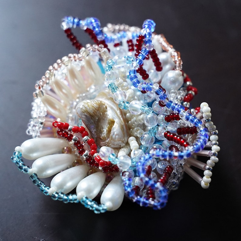 May include: A beaded brooch with a large iridescent shell, surrounded by white pearls, red, blue, and clear beads. The brooch is a cluster of beads in a variety of shapes and sizes.