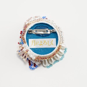 May include: A round, beaded brooch with a blue center and a white, pink, and blue beaded border. The back of the brooch has a blue fabric backing with a white label that says "月にょろし".