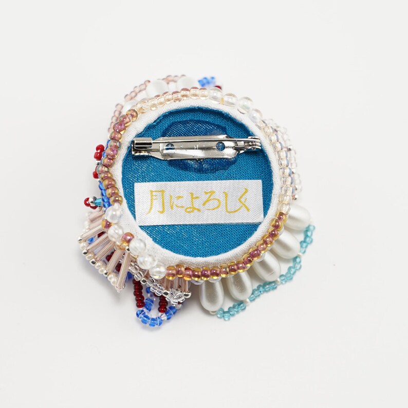 May include: A round, beaded brooch with a blue center and a white, pink, and blue beaded border. The back of the brooch has a blue fabric backing with a white label that says "月にょろし".