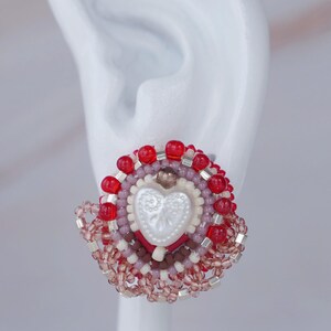 以下が含まれることがあります： 手作りのビーズイヤリング。中央に白いハートがあり、赤、ピンク、シルバーのビーズで囲まれています。円形のデザインは、詳細で手作りの外観をしています。イヤリングは白い背景に配置されています。