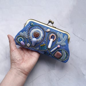 以下が含まれることがあります： 真鍮色の金属製キスロッククラスプが付いた青い布製のクラッチバッグ。外側には、複雑なビーズ細工、スパンコール、中央のハート、涙型のビーズ、滑らかな茶色の石など、さまざまな形のチャームが装飾されています。