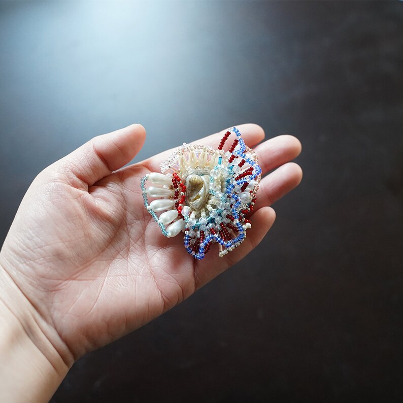 May include: A handmade brooch featuring a cluster of white pearls, blue and red beads, and a shell-like centerpiece. The brooch is a unique and intricate piece of jewelry.