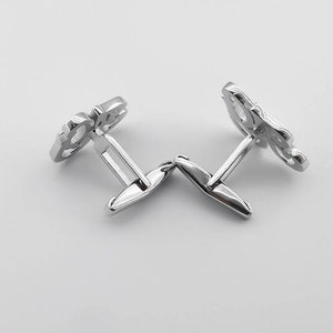 May include: A pair of silver cufflinks with a decorative design. The cufflinks are connected by a hinge.