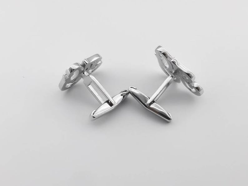 May include: A pair of silver cufflinks with a decorative design. The cufflinks are connected by a hinge.
