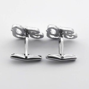 May include: A pair of silver cufflinks with the letters "DE" in a stylized font.